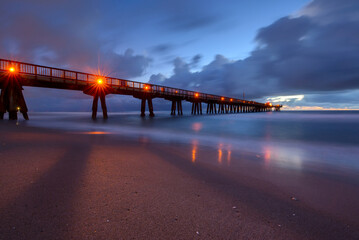 Long exposure of a calm beach and pier at dusk with glowing lights and reflective water