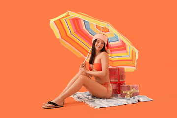 Young woman in Santa hat with beach umbrella and gifts sitting on orange background. Christmas in July