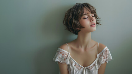 Studiio portrait of a young female model with short brown hair and posing in three-quarter profile with closed eyes. She is wearing a white lace top..