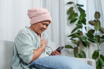 Elderly Asian female in warm headwear sits with hand on heart, phone aside,  Woman in pink...