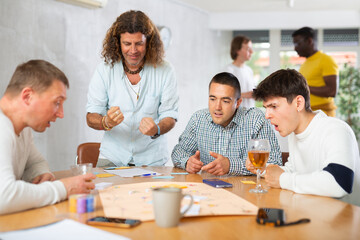 Expressive cheerful man with long wavy hair enthusiastically playing board game with male friends...