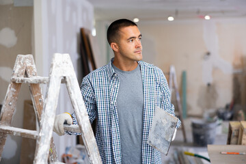 Portrait of man with float trowel standing at stepladder in apartment during repair works.