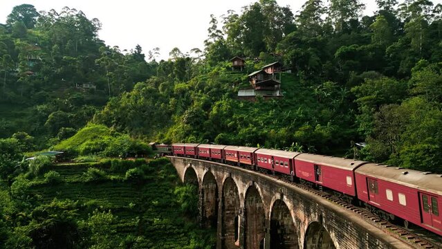 Sunrise illuminates Sri Lankan train crossing iconic Nine Arch Bridge surrounded by lush greenery. Red carriages glide over historic viaduct in hill country, Ella, scenic route for travelers.