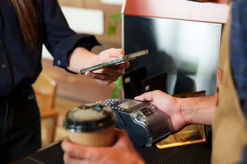 Barista processes mobile payment, customer holding smartphone over POS in daylight. Warmly lit coffee house scene with male attendant taking contactless payment from patron's smartphone at register.