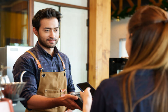 Smiling young male in checkered shirt and apron, engaged in friendly service at bakery counter. Joyful bakery worker in casual attire offers customer service, standing behind display shelf filled