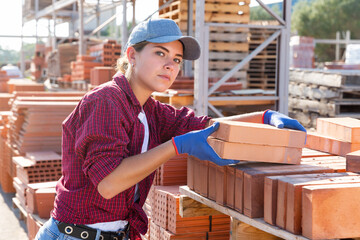 Female worker putting clay bricks on brick stack in outdoor construction material storage.