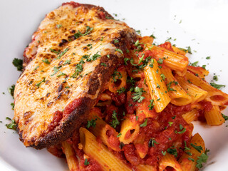 A cheese covered veal parmigiana on a bed of penne pasta with vegetable garnish on a white table