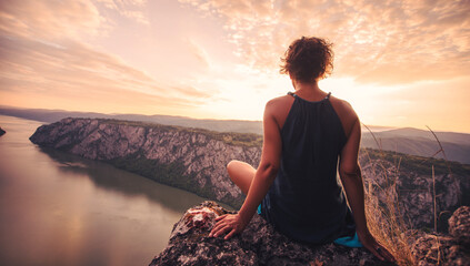 Person meditating on a cliff overlooking a tranquil river and mountains at sunset