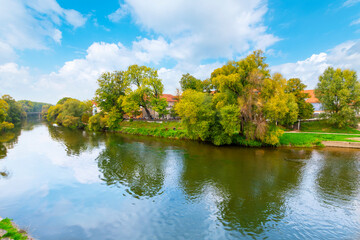 Colorful trees line the Danube tributary riverbank at the small island of Stadtamhof, with the medieval old town seen through the trees in Regensburg, Germany.