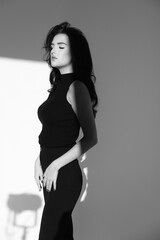 Fototapeta premium Brunette girl with closed eyes in black clothes on the background of a white wall. geometric shadows. Sensual black and white portraits of a model