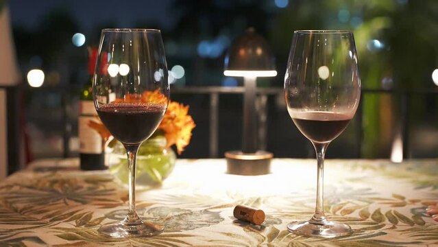 Close up motion of two red wine glasses on a table illuminated by lamp. Romantic dinner on a date in outdoor restaurant at night 
