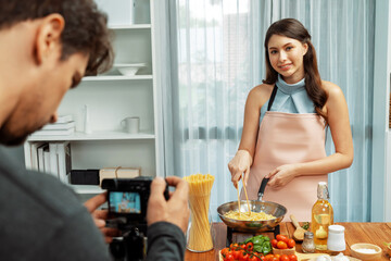 Cameraman recording to woman in chef influencer host cooking spaghetti with meat topped tomato sauce surrounded ingredients recipe, presenting special dish healthy food at modern studio. Postulate.