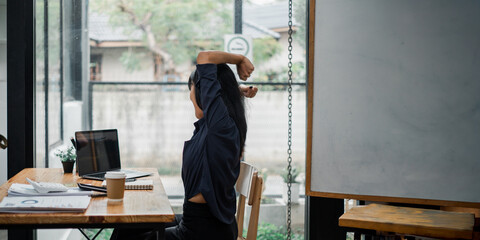 Back view of a female employee performing a stretch at her desk in a casual office environment.