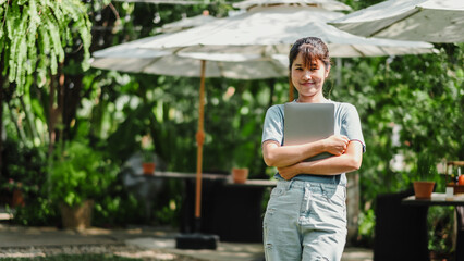 Female freelancer stands with her laptop in a lush garden, representing a serene remote work setting.