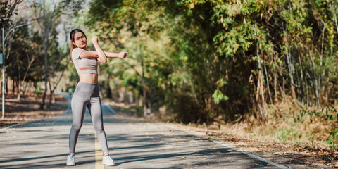 Fitness-focused woman engages in a stretching exercise for her arms while standing on a tree-lined path.