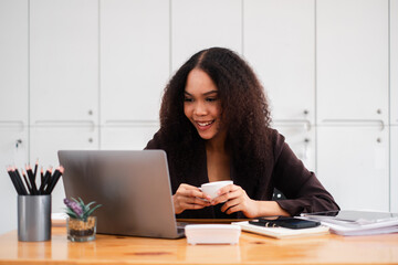 Smiling businesswoman enjoys a cup of coffee while working on her laptop, exuding a relaxed yet productive office atmosphere.