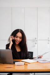Confident professional woman engages in a phone conversation while reviewing documents, managing her time effectively at her workspace.