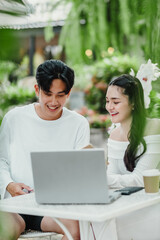 Attentive man and woman work together on a laptop, surrounded by the lush greenery of an outdoor garden cafe setting.