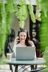 Smiling young woman using laptop and talking on phone in a green outdoor setting with coffee.