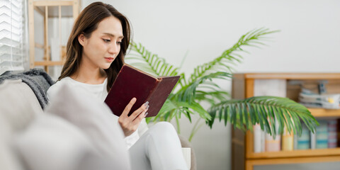 Young woman reading a book comfortably on a couch in a well-lit room with houseplant decorations.
