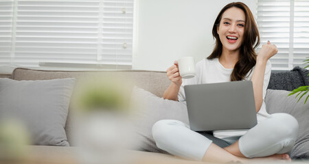 Joyful woman holding a coffee mug and making a fist pump gesture while using a laptop in a bright home setting.