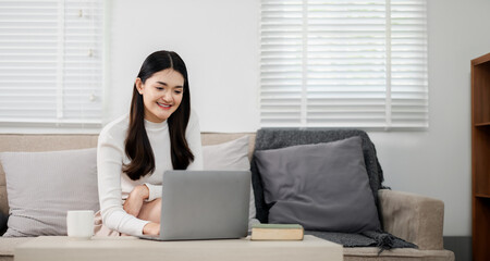 Contented woman enjoys working on her laptop, seated comfortably in a well-lit, tidy living room.