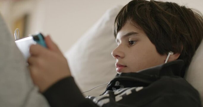 Cute young boy wearing headphones playing a game on a handheld gaming console, comfortably reclining on a couch indoors, daylight.