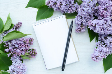 Blank notepad with pen and lilac flowers on white wooden background, top view, flat lay, copy space