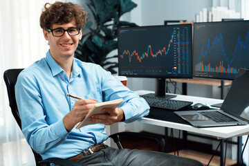 Profile of smiling young stock trader looking at camera, holding notepad and pen against on...