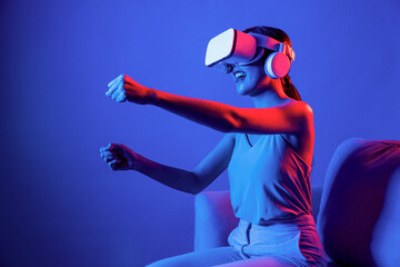 Smart Female sitting on sofa surrounded by neon light wear VR headset connecting metaverse, futuristic cyberspace community technology. Elegant woman play car racing game of meta world. Hallucination.