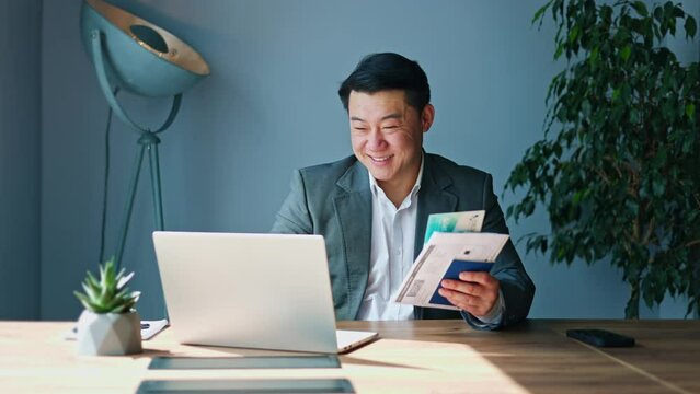 Smiling chinese worker typing on portable laptop and looking on printed tickets and documents in cabinet environment. Happy handsome male finishing online job and preparing to business trip indoors.