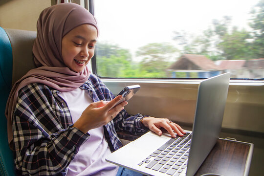 happy asian muslim woman holding mobile phone and sitting in front of laptop computer while sitting beside big window of train wagon. business traveling concept - Powered by Adobe