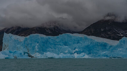 A block of blue ice rises like a wall above a glacial lake. Coastal mountains in clouds and fog. Icebergs float in glacial water. Perito moreno glacier. El Calafate. Argentina. Lago Argentino. 