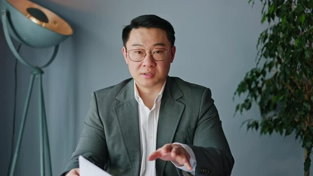 View from modern laptop camera of talkative japanese male gesturing while holding paper documents on background of designer cabinet interior. Handsome worker taking part in online conversation.
