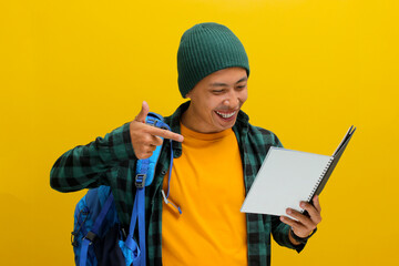 A smiling young Asian student, dressed in a beanie hat and casual shirt, carrying a backpack, points his finger to a notebook in his hand while standing against a yellow background