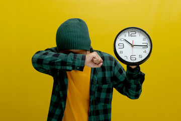 A tired young Asian man, dressed in a beanie hat and casual shirt, yawns while holding a clock,...
