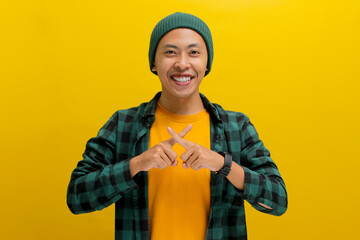 A young Asian man, dressed in a beanie hat and a casual shirt, is making an X sign gesture by...