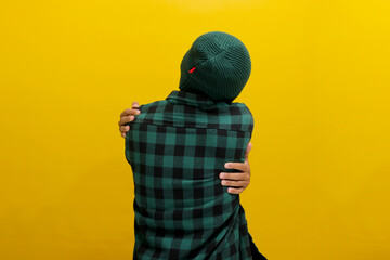 Back view of Young Asian man, wearing a beanie hat and casual shirt, hugs himself, symbolizing the...