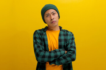 Bored young Asian man wearing a beanie hat and casual outfit keeping arms folded, frowning his face in displeasure while standing against yellow background.