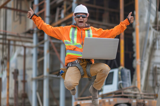 A happy construction worker jumping in the air with his white helmet, wearing a high visibility safety jacket and holding a laptop computer under one arm, with black sunglasses on his face.