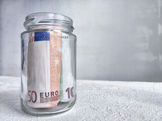 Glass Jar Filled With Euro Banknotes on a Textured White Surface. Euro currency in clear jar as...