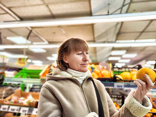 Middle-Aged Woman Holding an Orange in a Grocery Store. Woman examines an orange, considering nutrition in a store aisle. Concept of proper nutrition