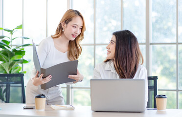 Asian young pretty female businesswoman team smiling discussing project on laptop standing and sitting at table in office. Two entrepreneurs colleagues of professional business people working together