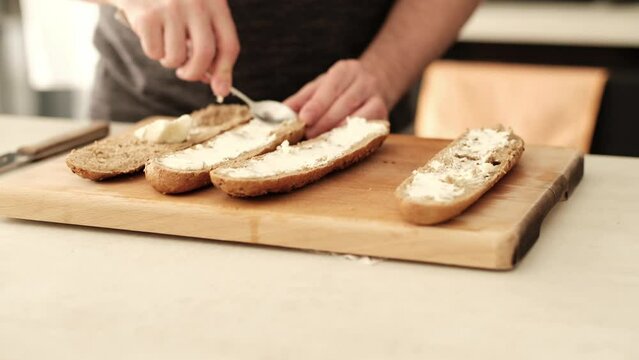 Caucasian male spreading creamy cheese on brown flour baguette on the kitchen countertop.