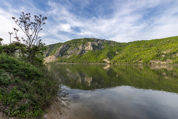  serene lakeside scene with a prominent mountain cliff in the background. The tranquil water reflects the greenery and the sky, creating a picturesque setting.