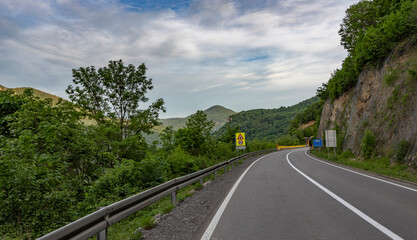 Fototapeta premium A winding mountain road with a guardrail on the left, bordered by lush green trees and vegetation. Yellow caution signs and white road markings indicate a driving perspective on an overcast day