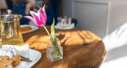 A pink tulip in a glass vase on a wooden table, accompanied by a tea set and gingerbread cookies on...