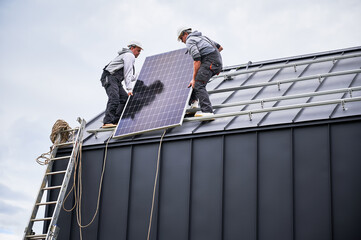 Electricians installing solar panel system on roof of house. Men workers in helmets carrying...