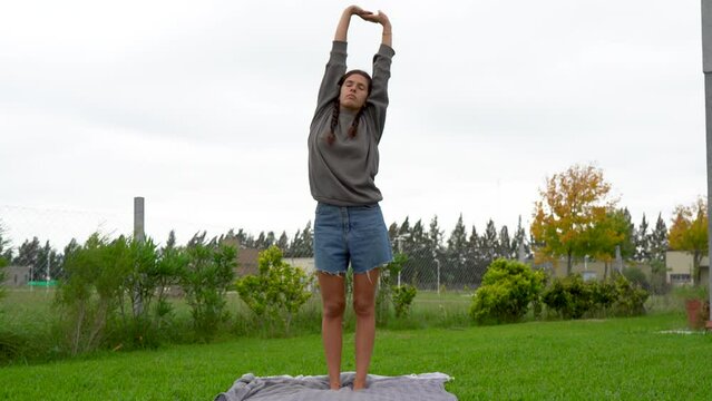 Young woman stands and stretches on blanket on lawn on cloudy day, full shot
