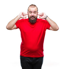 Young caucasian hipster man wearing red shirt over isolated background covering ears with fingers with annoyed expression for the noise of loud music. Deaf concept.